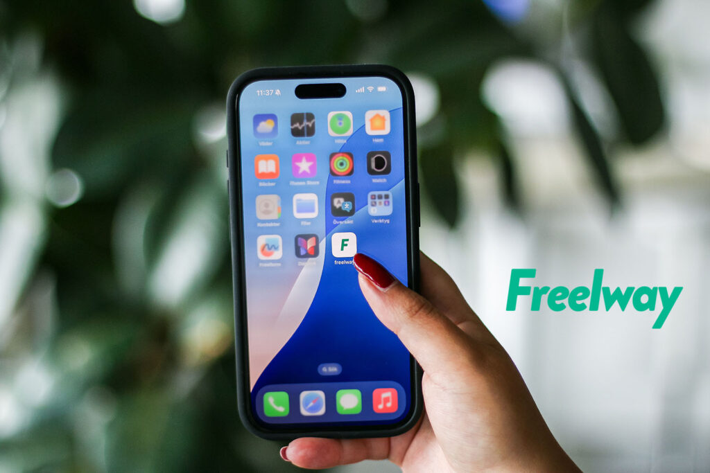 A hand with thumb poised to tap the Freelway app on a smartphone, with green leaves blurred in the background.