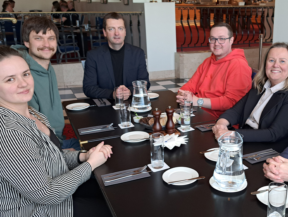 Five people sitting and smiling around a dinner table set with glasses and carafes in a restaurant.