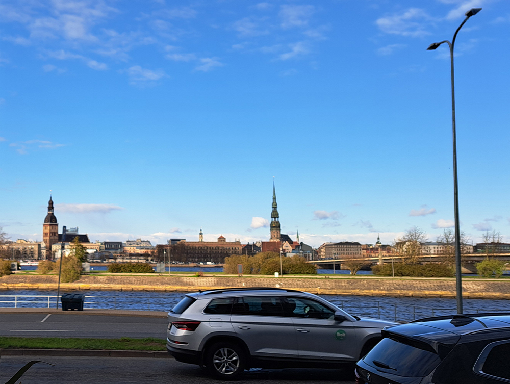 A city skyline with historic church spires across a river under a blue sky, with a silver car in the foreground.