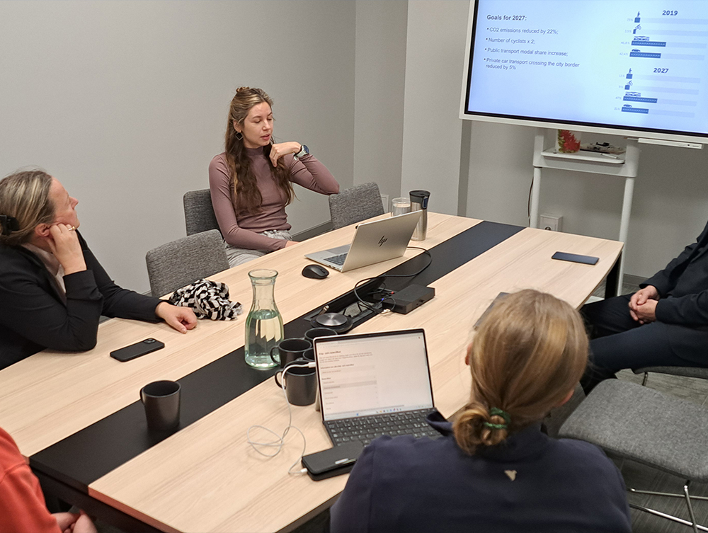 A woman presenting at a conference table next to a screen showing "Goals for 2027" charts.