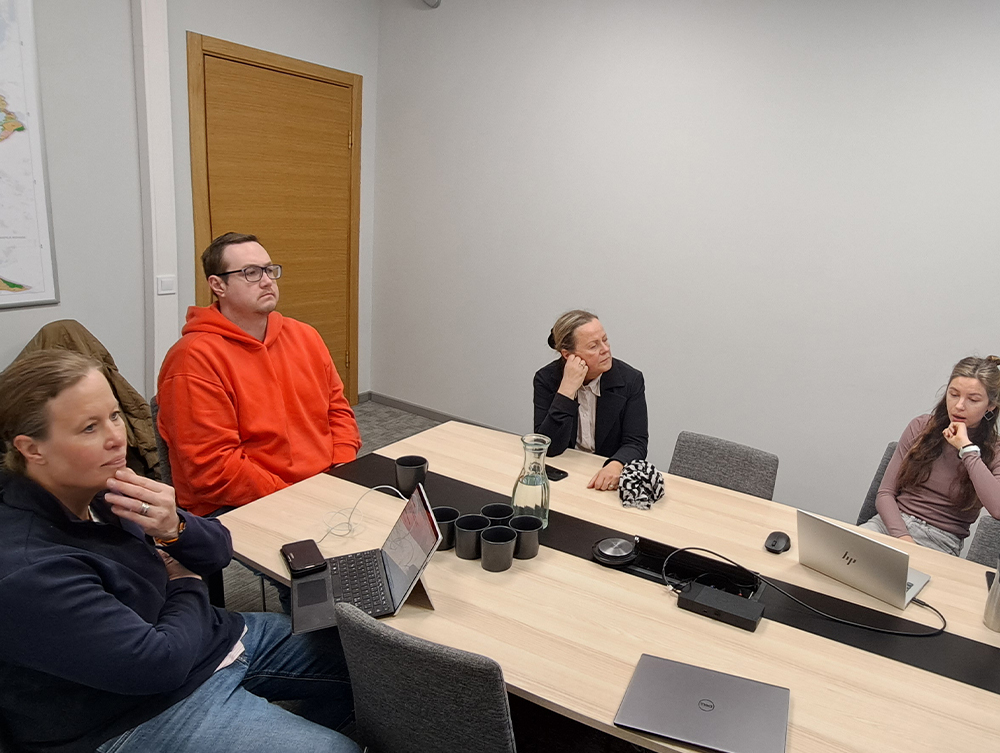 People sitting around a meeting room table with laptops, listening attentively to a presentation.
