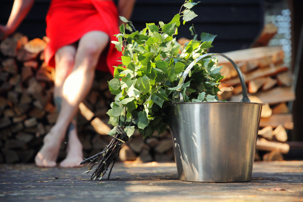 A person sitting outside after sauna. In the front of the photo there is a Sauna bucket and a sauna broom make of a birch branch.