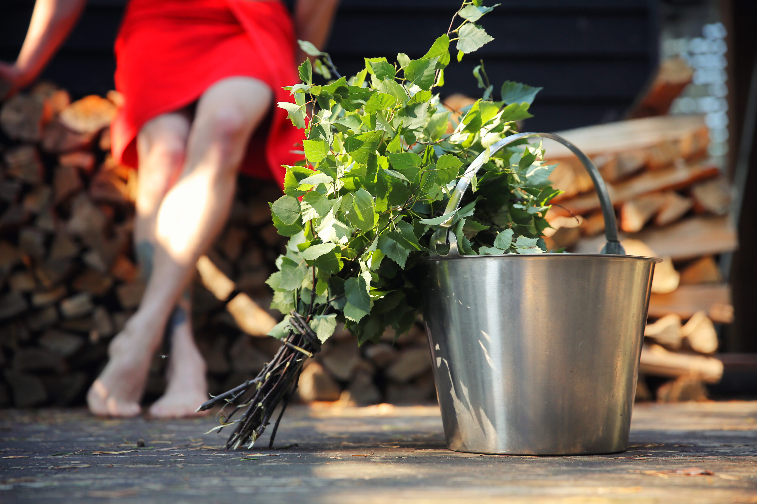 A person sitting outside after sauna. In the front of the photo there is a Sauna bucket and a sauna broom make of a birch branch.