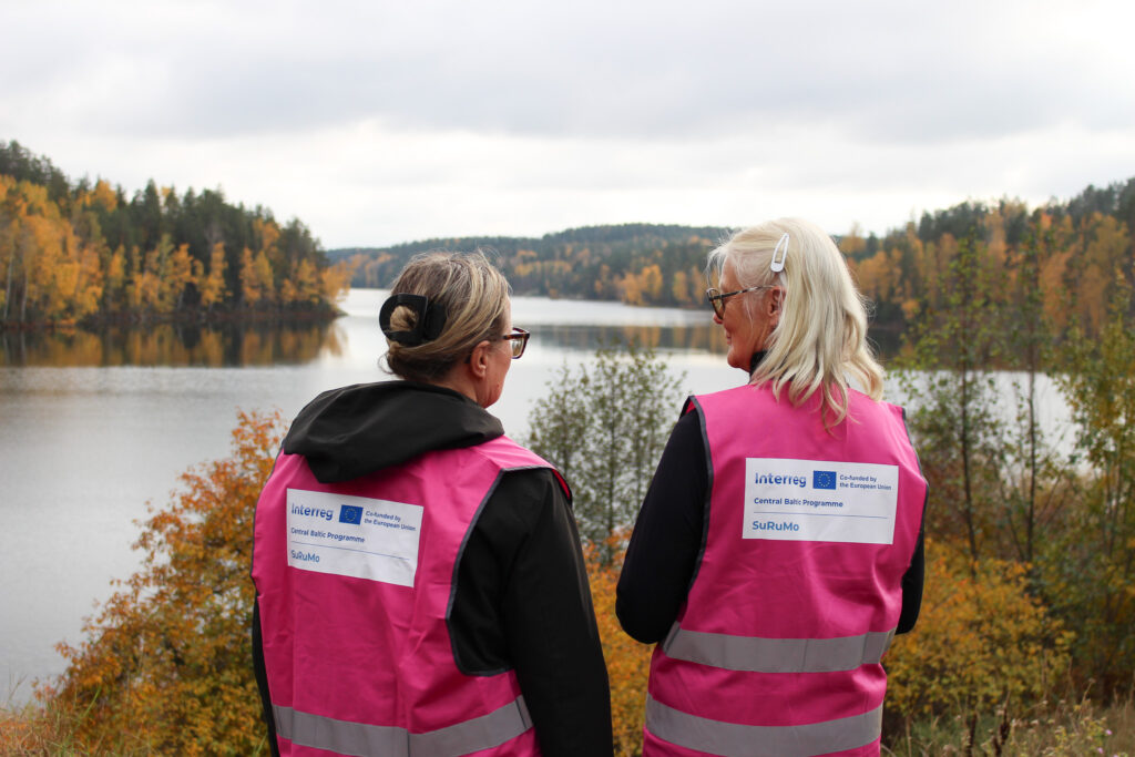 Two women in pink SuRuMo-vests stand by a lake, looking out over autumn forest and water.