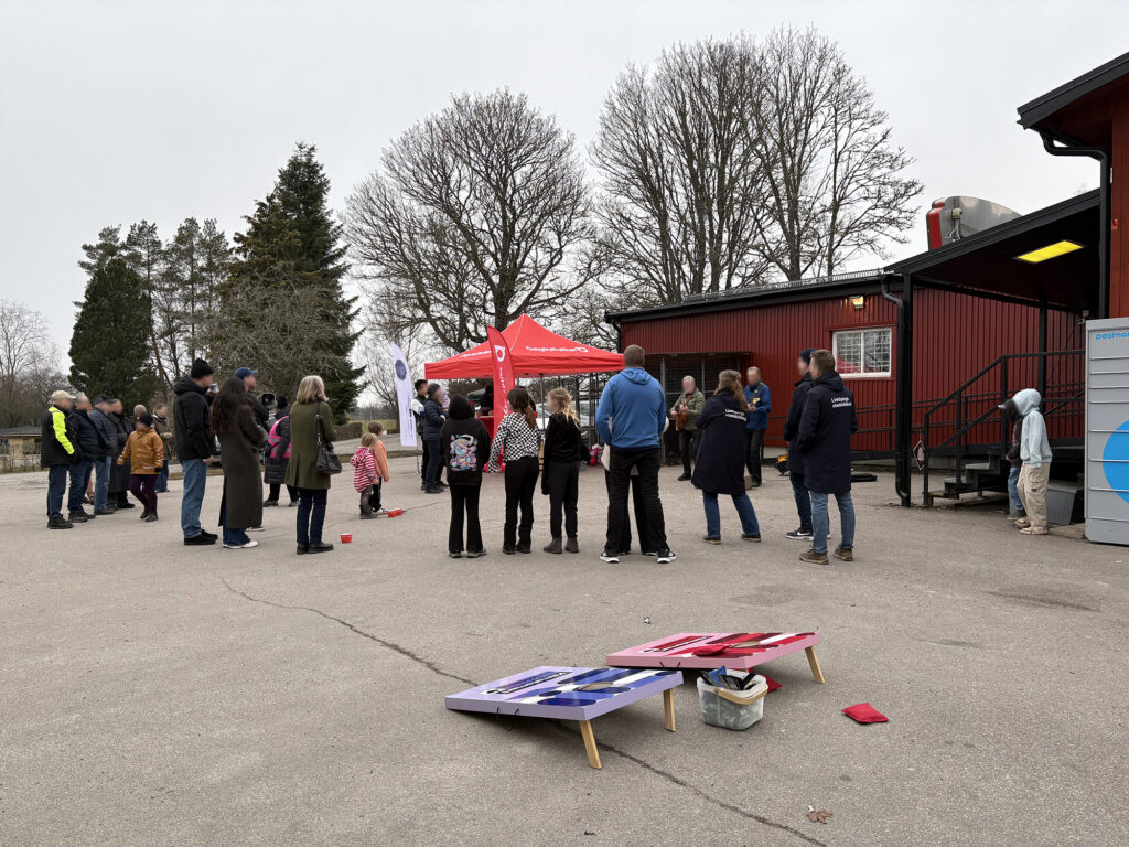 An outdoor gathering in front of a red building with people watching a performance near a red tent, with a cornhole game in the foreground.