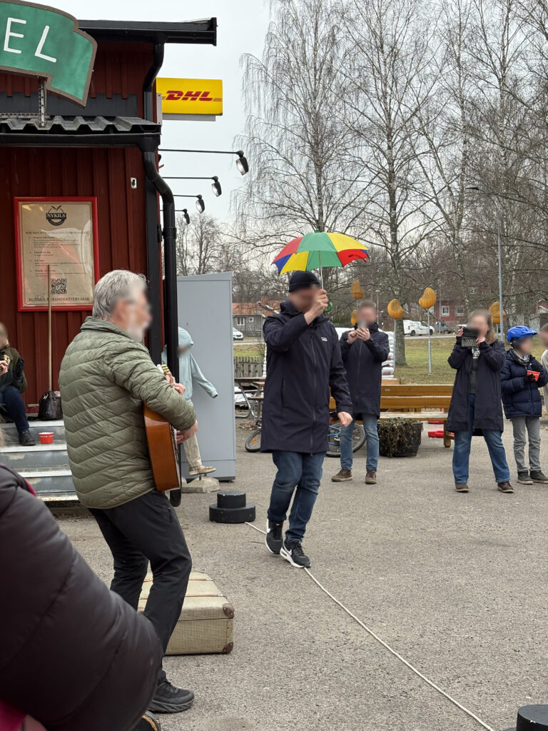 A man balancing on a tightrope while holding a colorful umbrella, as a guitarist plays music and onlookers watch