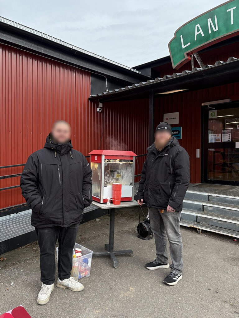 Two men standing outside a red building next to a steaming popcorn machine on a table.