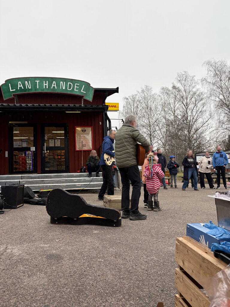 A live musical performance outside a red village store named "Lanthandel" with a small crowd gathered on the asphalt.