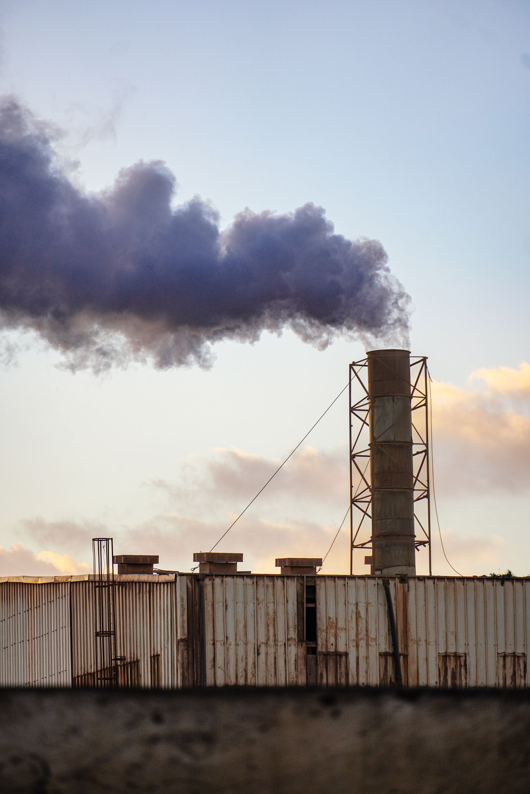 A factory chimney releasing smoke into the sky, silhouetted against a sunset. Environmental impact theme.
