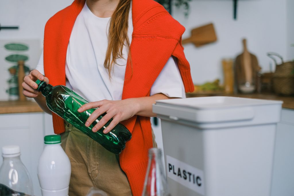 Crop anonymous female in casual clothes standing with plastic bottle while sorting garbage in light kitchen in daytime